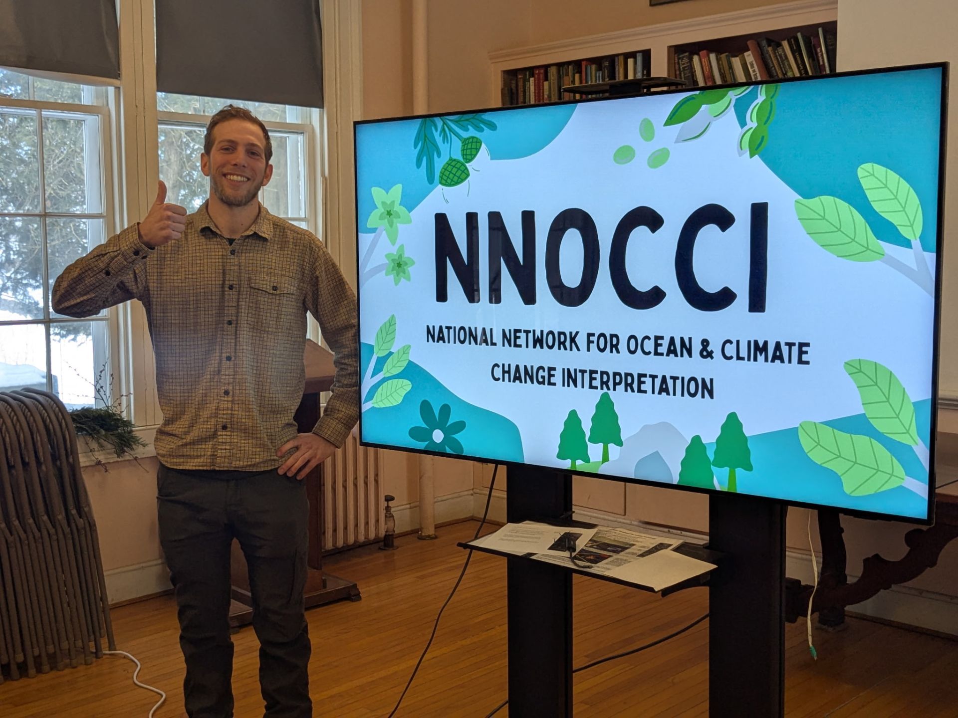 Jake in front of TV screen with NNOCCI slide
